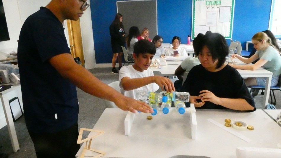 A team of three students testing their paper bridge by placing 5 full 500 ml water bottles on it